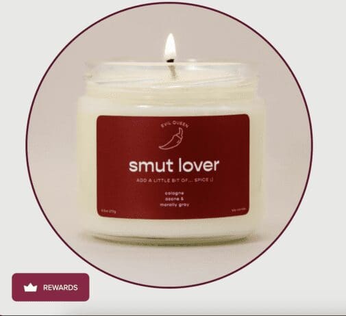 A lit candle in a clear glass jar with a maroon label that reads 'Smut Lover' in bold white text. Above the title is a small illustration of a chili pepper with the words 'Evil Queen' written above it. Below the title, the label says 'Add a little bit of... spice :)' and lists the scent notes as 'cologne, ozone & morally gray.' The candle is set against a neutral background, and a small rewards icon is visible in the lower-left corner of the image.